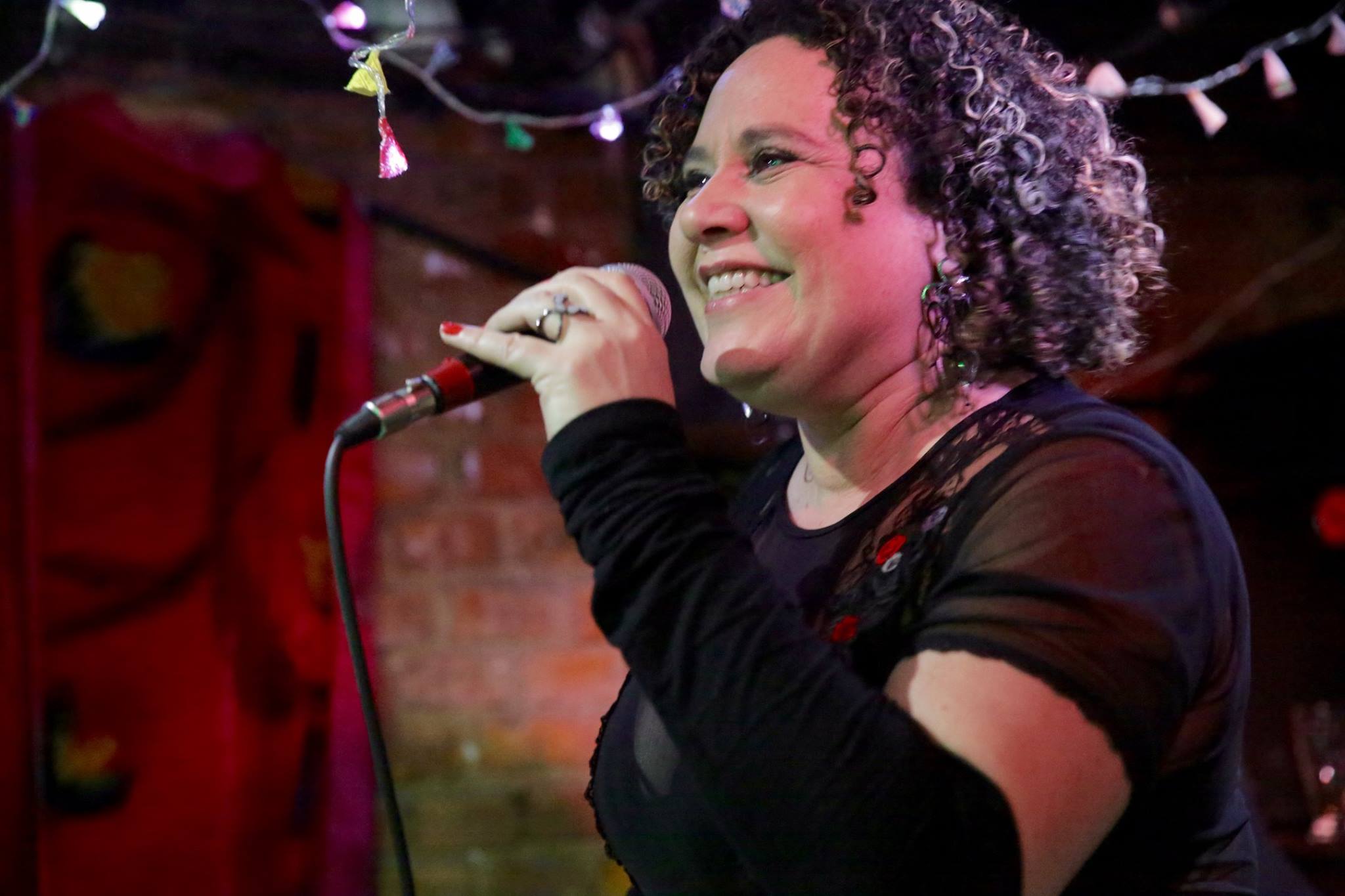 Cantora Lu Barros, canta com microfone na mão. Ela está sorridente, tem os cabelos cacheados e usa um vestido preto.