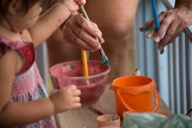 A imagem mostra mãos de uma criança e de uma mulher adulta mexendo com tintas naturais para pintar uma casinha de boneca.